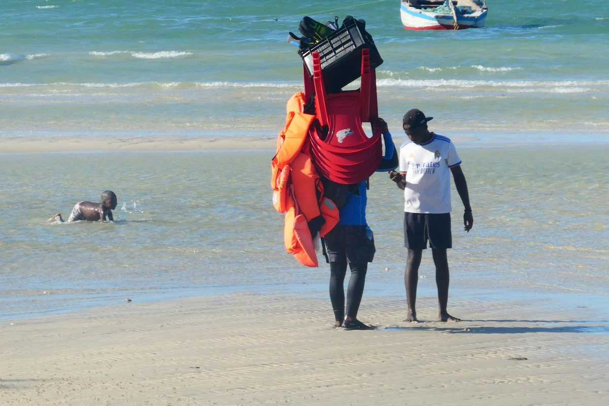 Mozambican man walking along the beach in Vilanculos carrying chairs, with a child playing in the clear blue Indian Ocean.