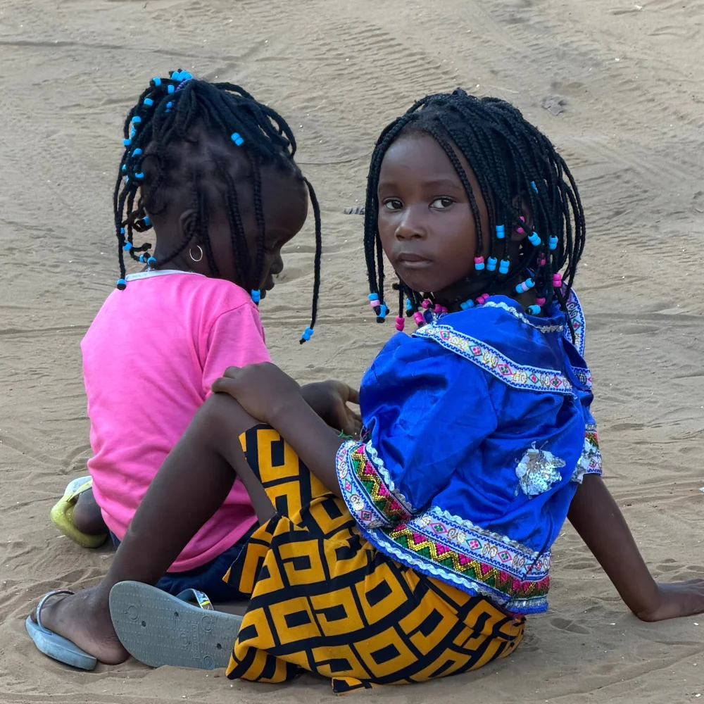 Kleurrijke Mozambikaanse meisjes Twee Mozambikaanse meisjes zitten samen op het zand, gekleed in kleurrijke kleding en met gevlochten haar versierd met kralen.